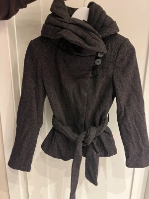Mörk brungrå kappa med stor krage från Zara - Brungrå kort kappa från Zara Basic med stor, svepande krage och knytbälte i midjan. Jackan har tre dekorativa knappar framtill och är figursydd för en schysst siluett. Perfekt för dig som gillar stilrena och moderna detaljer. Liten i storleken, känns som den är i storlek S.
