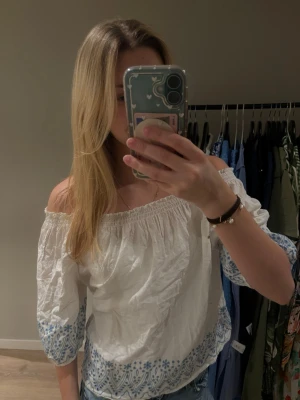 Vit offshoulder blus Amelie & Me - Superfin vit offshoulder blus från Amelie & Me med blå broderade blommor och mönster längs ärmslut och nederkant. Blusen har resår upptill och trekvartsärmar, perfekt till jeans eller kjol för en fräsch look.