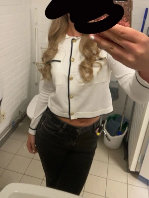 Vit croppad blus med guldknappar - Snygg vit croppad blus med svarta detaljer och guldiga knappar framtill. Långa ärmar med svarta kanter och två dekorativa fickor på bröstet. Använd en gång