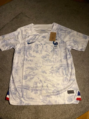 Nike Frankrike fotbollströja M - Säljer en officiell Nike Frankrike fotbollströja i storlek M. Tröjan är vit med ljusblått mönster av franska landmärken och har broderad tupp-logga och Nike swoosh i blått. Tillverkad i Dri-FIT polyester för bästa komfort. Trikolor-detalj i nederkant.
