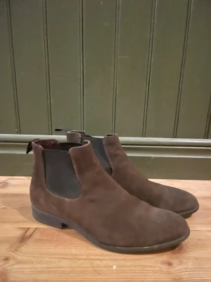 Bruna chelsea boots vaga bond  - Säljer ett par stilrena bruna chelsea boots i mjuk mocka. Skorna har elastiska partier på sidorna och en låg klack. Perfekt för kyligare dagar! Nypris ligger på 1800 kr, sälj här för 50% av nypriset🤩 skicket är 7,5/10 