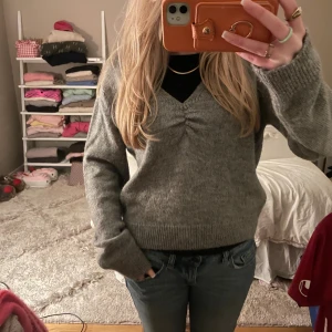 Grå v-ringad stickad tröja JDY - Mysig grå stickad tröja från JDY med v-ringning och snygg rynkdetalj framtill. Långärmad modell med ribbade muddar och avslappnad passform. Perfekt att slänga på sig när det är kyligt ute.