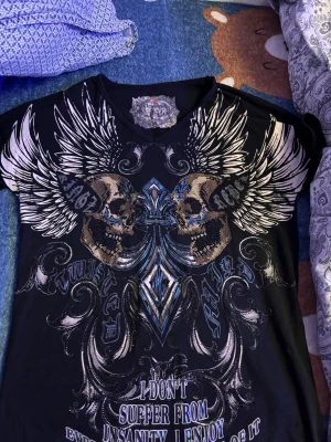 Svart t-shirt med dödskallar från Kingz - Cool svart t-shirt från Kingz med stort tryck av dödskallar, vingar och blå detaljer på framsidan. T-shirten har v-ringning och korta ärmar. Materialet är mjuk bomull med lite elastan för extra stretch. Grym statement-piece för dig som gillar unika prints.