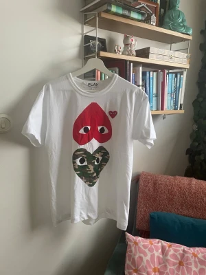 Comme des Garçons Play x Bape T-shirt - Säljer en vit Comme des Garçons Play x Bape t-shirt i storlek M. Framsidan har två stora hjärtan, ett rött och ett i camouflage, båda med ögon. Klassisk rund hals och korta ärmar. Tillverkad i 100% bomull för skön känsla. Tröjan är nästan oanvänd. Vid minsta fråga är det bara höra av sig!