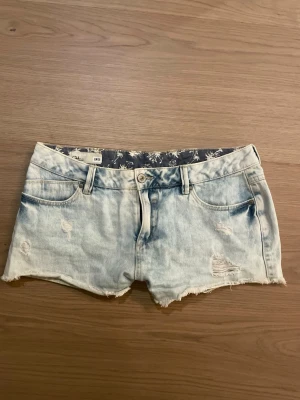 Ljusblå lågmidjade jeansshorts st S/M - Säljer ett par ljusblå jeansshorts med råa kanter och slitna detaljer. Klassisk femficksmodell med knapp och dragkedja framtill. Perfekta till sommaren och ger en avslappnad vibe. Midjemått på cirka 40-41 cm