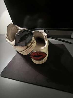 Vita sneakers med läppdetalj bak - Unika vita sneakers i skinn med snörning och coola läppar i blått och rött bak på hälen. Skorna har platt sula och rund tå, och detaljer i metall på läpparna. Perfekt för dig som vill sticka ut med din stil.