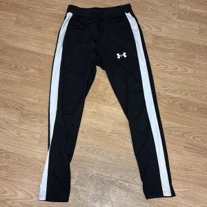 Svarta träningsbyxor från Under Armour - Svarta träningsbyxor från Under Armour med vita ränder längs sidorna och logga på låret. Byxorna har smal passform, elastisk midja med snörning och är gjorda i ett stretchigt syntetmaterial som passar perfekt för sport och träning. Hör av er vid intresse!