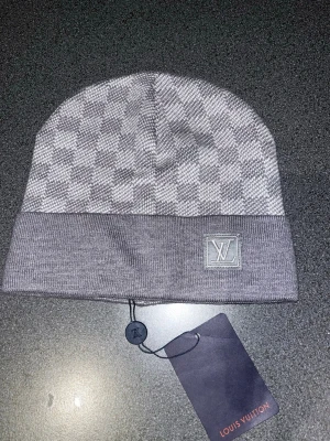 Grå mössa med rutigt mönster - Säljer en snygg grå mössa med klassiskt rutigt mönster. Mössan är stickad och har en bred uppvikt kant. Perfekt accessoar för dig som vill ha en stilren och exklusiv look.