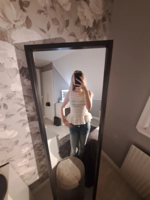 Vit volangtop från H&M Divided XS - Säljer en söt vit topp från H&M Divided i storlek XS. Toppen är ärmlös med volang nertill och har ett ribbat, lätt transparent tyg med horisontella ränder. Perfekt till jeans eller kjol för en fräsch look.