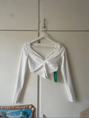 Vit croppad topp med knyt från SHEIN - Snygg vit croppad topp från SHEIN med långa ärmar och v-ringning. Toppen har ett trendigt knyt framtill och är tillverkad i ett mjukt bomullsliknande material. Aldrig använt den eftersom den inte är min stil.