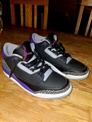 Nike Air Jordan 3 Court Purple 44.5 - Nike Air Jordan 3 Court Purple i svart skinn med lila och orange detaljer. Klassisk elephant print runt tå och häl, samt vit och grå sula. Skorna har snörning och rund tå. Insidan är fodrad i orange textil och modellen har ikonisk Jumpman-logga på plösen.