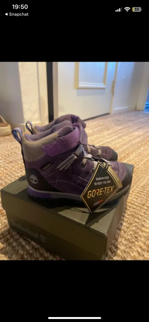 Lila Timberland kängor barn strl 24 - Säljer ett par lila Timberland kängor för barn i storlek 24. Skorna har Gore-Tex-membran, grov sula och kardborreband för enkel påtagning. Tillverkade i mocka och syntet, perfekta för äventyr utomhus. Färg: mörklila och grå.