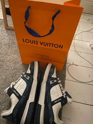 Louis Vuitton blåvita sneakers - Säljer ett par Louis Vuitton sneakers i blått och vitt med klassiskt monogrammönster. Skorna har snörning, rund tå och platt sula. Tillverkade i skinn och textil med detaljer i mörkblått och vita LV-loggor på sidan och tungan. Snygg och exklusiv modell för dig som gillar streetstyle.