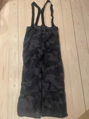 Svarta camoflash skid/överdragsbyxor - Säljer ett par svarta täckbyxor från Everest i storlek 152. Byxorna har ett mörkt camomönster, justerbara hängslen och dragkedja fram. Perfekta för vintern och outdoor-aktiviteter. Dom är lite slitna som ni ser på bilderna men inget så är hål igenom!