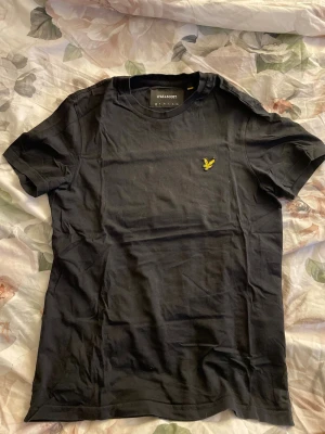 Svart t-shirt från Lyle & Scott - Klassisk svart t-shirt från Lyle & Scott med gul broderad logga på bröstet. T-shirten har rund halsringning och korta ärmar. Inga skador eller fläckar på tröjan, säljs pågrund utav att den aldrig kommer till användning. 