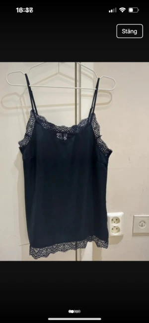 Marinblå linnetopp med spetsdetaljer från Vila  - Snygg  linnetopp med tunna axelband och dekorativ spets längs halsringning och nederkant. Toppen har en mjuk, slät yta och är lätt och luftig, perfekt för att styla med jeans eller kjol. Spetsen ger en extra touch av elegans och gör toppen superfin.