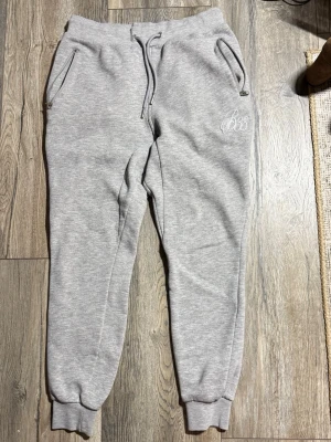 Grå mjukisbyxor från Bee Inspired - Snygga ljusgrå mjukisbyxor från Bee Inspired med broderad logga på vänster ficka. Byxorna har dragsko i midjan, muddar vid bensluten och dragkedjor på fickorna. Perfekta för chill eller träning.