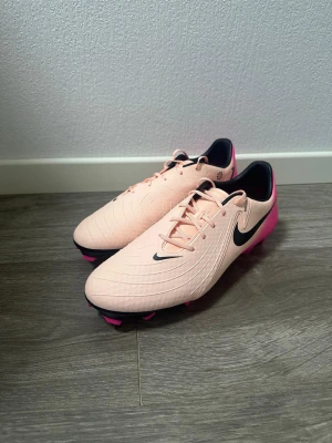 Fotbollsskor i ljusrosa och rosa - Säljer ett par fotbollsskor i en snygg ljusrosa färg med starkt rosa detaljer på hälen och sulan. Skorna har svart logga, snörning och rund tå. Ovandelen är i syntetmaterial med mönstrade cirklar för extra grepp på bollen. Perfekta för dig som vill sticka ut på planen!