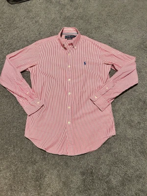 Rödvit randig skjorta från Ralph Lauren - Klassisk skjorta från Ralph Lauren i rödvitt randigt mönster. Skjortan har lång ärm, knappar hela vägen och blå broderad logga på bröstet. Classic fit-modell i mjuk bomull, perfekt till jeans eller chinos.