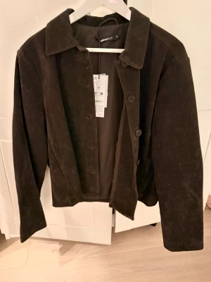 Mörkbrun mocka imitation från Gina Tricot - Säljer en stilren mörkbrun kort jacka  från Gina Tricot. Jackan har klassisk krage, raka ärmar och knäppning med stora knappar framtill. Nypris 699 kr 