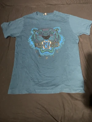 Blå Kenzo t-shirt med tigertryck - Säljer en blå t-shirt från Kenzo med ikoniskt tigertryck och texten 'Kenzo Paris' framtill. T-shirten har rund hals, korta ärmar och är gjord i mjuk bomull.