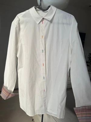 Vit skjorta från Paul Smith - Säljer en vit skjorta från Paul Smith med färgglada knapphål och klassisk krage. Manschetterna har snyggt randigt foder i flera färger som syns när du viker upp ärmarna. Skjortan är i bomull och har en clean, stilren look med en twist.