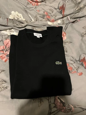 Svart stickad tröja från Lacoste - Klassisk svart stickad tröja från Lacoste med rund halsringning och den ikoniska gröna krokodilloggan på bröstet. Regular fit och ribbade muddar vid hals, ärmslut och nederkant. Perfekt till en clean och stilren look.