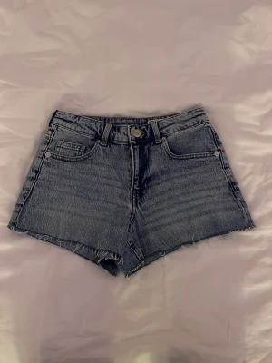 Jeansshorts  - Säljer ett par klassiska blå denimshorts från veromoda med fransiga benslut. Shortsen har fem fickor, bälteshällor och knappgylf. Perfekta till sommaren för en avslappnad och trendig look.
