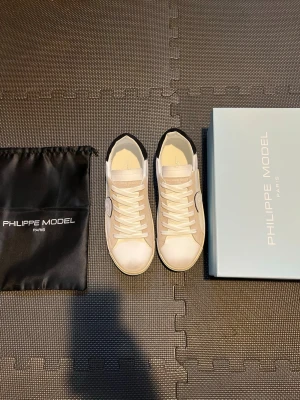 Philippe Model vita sneakers - Snygga vita sneakers från Philippe Model med beige mockadetaljer och svart häl. Skorna har klassisk snörning, rund tå och logotyp på sidan. Tillverkade i skinn och mocka för en lyxig känsla. Kommer med originalpåse och låda.
