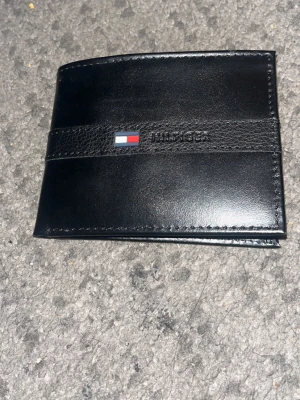 Svart läder plånbok från Tommy Hilfiger - Snygg svart plånbok från Tommy Hilfiger i läder med klassisk logga på framsidan. Insidan har flera kortfack och ett fack med röd- och marinblått randigt tyg. Perfekt för dig som vill ha en stilren och praktisk accessoar. Helt enkelt en väldigt praktiskt plånbok.