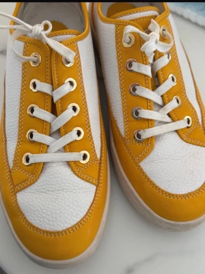 Vita och gula sneakers från Michael Kors - Säljer ett par snygga sneakers från Michael Kors i vitt och gult skinn. Skorna har vita snören, guldfärgade öljetter och en cool kontrast mellan det släta och det strukturerade lädret. Perfekta för dig som vill sticka ut med färg och stil.