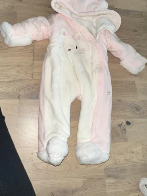 Söt teddyoverall med kaninmotiv Rosa overall - Mysig teddyoverall i ljusrosa och vitt med gulligt kaninansikte och öron på magen. Overallen har huva, långa ärmar och fluffiga muddar vid ärmar och ben. Perfekt för kalla dagar och supergosig att ha på sig.Söt rosa overall med en kanin design på framsidan och en huva med små öron.