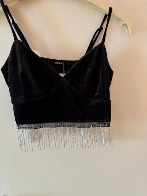 Svart festlig topp från BikBok  - Snygg festlig topp från BikBok i mjuk sammet. Toppen har tunna axelband, v-ringning och glittriga strassfransar längs nederkanten. Helt ny och oanvänd.