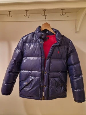 Blå pufferjacka från Polo Ralph Lauren - Snygg blå pufferjacka från Polo Ralph Lauren med röd insida och klassisk logga på bröstet. Jackan har hög krage, huva och stora fickor med knappar. Perfekt för kalla dagar och riktigt skön att bära.