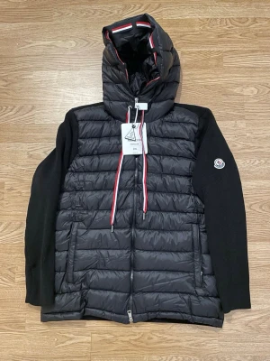 Svart Moncler cardigan  2XL passar M/L - Säljer en svart pufferjacka från Moncler i storlek 2XL. Jackan har stickade ärmar, quiltad kropp och huva med röd, vit och blå detalj. Klassisk Moncler-logga på ärmen och dragkedja framtill. Perfekt för dig som vill ha en snygg och varm jacka. Passar M/L