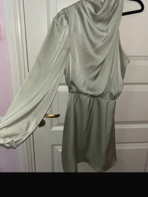 Ljusgrön satin one shoulder klänning - Säljer en ljusgrön kortklänning i satin med en ärm och oneshoulder-design. Klänningen har lös passform i midjan, draperad halsringning och stängs med dragkedja och knappar bak i nacken. Perfekt för dig som vill sticka ut med en unik look.