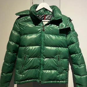 Grön Maya pufferjacka från Moncler - Säljer en grön pufferjacka från Moncler med hög krage och avtagbar huva. Jackan har dragkedja framtill, två fickor med dragkedja och en patch med Moncler-logga på ärmen. Perfekt för kalla dagar. Köpt på Merchsweden för drygt ett år sedan. 