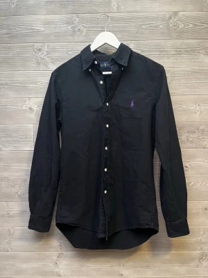 Svart skjorta från Ralph Lauren S - Klassisk svart skjorta från Ralph Lauren i storlek S. Skjortan har lång ärm, knappar framtill och en lila broderad logga på bröstet. Tillverkad i bomull för en skön känsla och clean look.