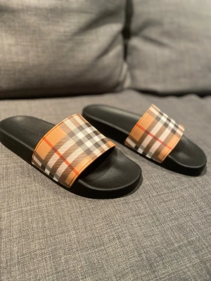 Burberry tofflor  - Säljer ett par Burberry sandaler med svart sula och bred rem i det ikoniska beige, bruna och röda rutmönstret. Tillverkade i syntetmaterial med präglad Burberry-logga på sidan och sulan. Perfekta för sommaren
