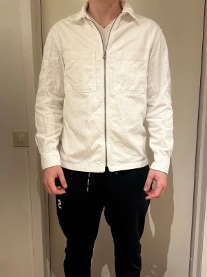Vit overshirt från H&M med dragkedja - Säljer en vit overshirt från H&M i relaxed fit. Jackan har två bröstfickor, hel dragkedja framtill och klassisk krage. Tillverkad i bomull och passar perfekt för lager på lager. Enkel och stilren design som funkar till det mesta.