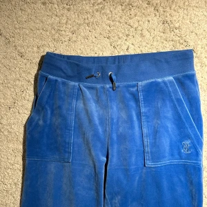 Blåa mjukisbyxor Juicy Couture S - Blåa mjukisbyxor från Juicy Couture i mjuk sammet med bred resårmidja och snörning. Byxorna har stora fickor fram och bakfickor med knappar samt broderad JC-logga på ena benet. 