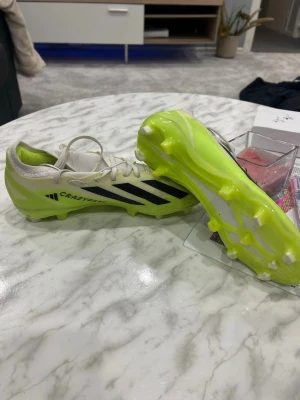 Adidas Crazyfast fotbollsskor 39⅓ - Säljer ett par Adidas Crazyfast fotbollsskor i vitt och neongult med svarta ränder. Skorna har snörning och en låg profil med platt sula och dobbar för gräsplan. Tillverkade i syntetmaterial för lätt känsla och maxad speed på planen.