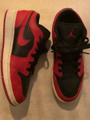 Nike Air Jordan 1 Low Svart/Röd - Säljer ett par Nike Air Jordan 1 Low i svart och rött skinn med klassisk Jumpman-logga på plösen. Skorna har svart snörning, perforerad tå och vit sula med röd yttersula. Perfekta för dig som vill ha en ikonisk sneaker med streetstyle-vibe.