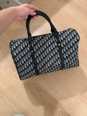 Dior monogram axelväska i textil - Säljer en snygg Dior axelväska med klassiskt monogrammönster i svart och vitt. Väskan har svarta detaljer i skinn, guldfärgade metalldelar och en bred axelrem med Dior-logga. Insidan är fodrad i rött tyg och har en innerficka. Perfekt för dig som gillar statement-accessoarer.