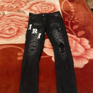 Svarta jeans med slitningar från Amiri - Snygga svarta jeans från Amiri med coola slitningar och broderade bokstäver. Perfekta för en edgy look. Jeansen har en klassisk femficksdesign och knappgylf. Storlek S men funkar som storlek M