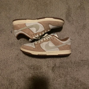 Nike Dunk - Medium Curry - Storlek 45 - Nike Dunk Low sneakers i beige och ljusbrun mocka med vita detaljer och snörning. Skorna har en klassisk låg siluett, rund tå och vit sula med bruna partier. Swoosh-logga på sidan och broderad Nike-text bak. Perfekta för dig som gillar stilrena och neutrala färger. Storlek 45. Knappt använda kanske 6-7 gånger
