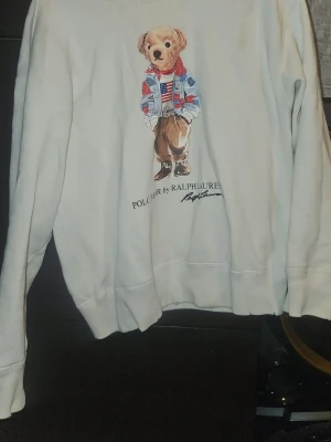 Vit sweatshirt Polo Bear Ralph Lauren - Vit sweatshirt från Polo Ralph Lauren med ikoniskt Polo Bear-tryck framtill. Tröjan har rund hals, långa ärmar och ribbade muddar. Perfekt för dig som gillar klassisk streetwear med en twist.