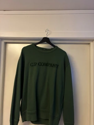 Grön sweatshirt från C.P. Company - Säljer en grön sweatshirt från C.P. Company med broderad logga på bröstet. Tröjan har rund hals, lång ärm och är gjord i mjuk bomull. Perfekt för dig som gillar stilrena och bekväma plagg.