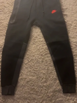 Svarta Nike mjukisbyxor med röd logga - Säljer ett par svarta mjukisbyxor från Nike med röd logga på vänster lår. Byxorna har snörning i midjan, muddar vid bensluten och dragkedja på ena fickan. Perfekta för chill eller träning. Skriv om du har några frågor.