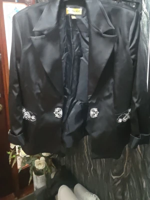 Svart kavaj med paljettbroderi - Säljer en svart kavaj från Polen Poe med glansig finish och stora slag. Kavajen har detaljerade paljettbroderier och pärlor på fickorna och ryggen, vilket ger en unik och lyxig vibe. Perfekt för dig som vill sticka ut med coola detaljer och elegant look.